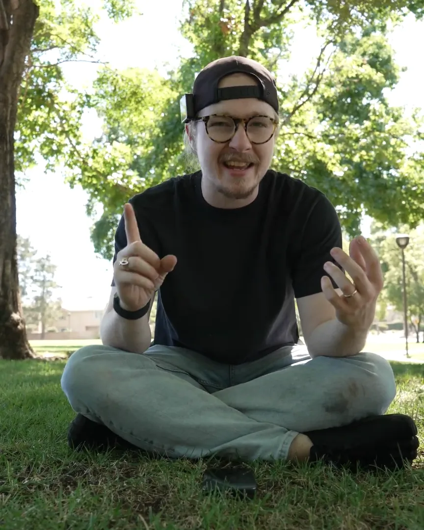 David sitting cross-legged in the park, smiling and pointing one finger up